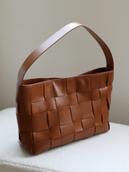 Angled view of the classic brown genuine leather woven tote bag resting on a soft surface, showcasing its elegant wide strap and structured silhouette.