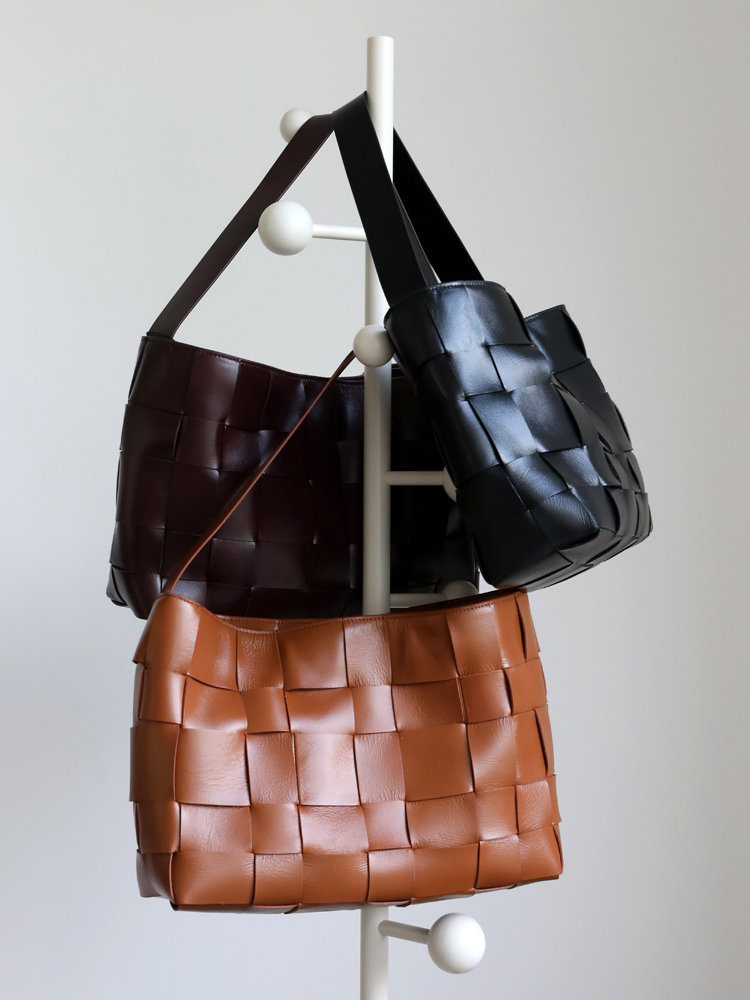 A stylish display of three genuine leather wide-weave shoulder bags in classic brown, black, and dark burgundy, hanging on a minimalist rack.