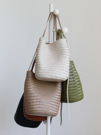 A stylish display of handwoven vegan leather bucket tote bags hanging on a rack, featuring cream, khaki, olive green, black, and brown colorways.