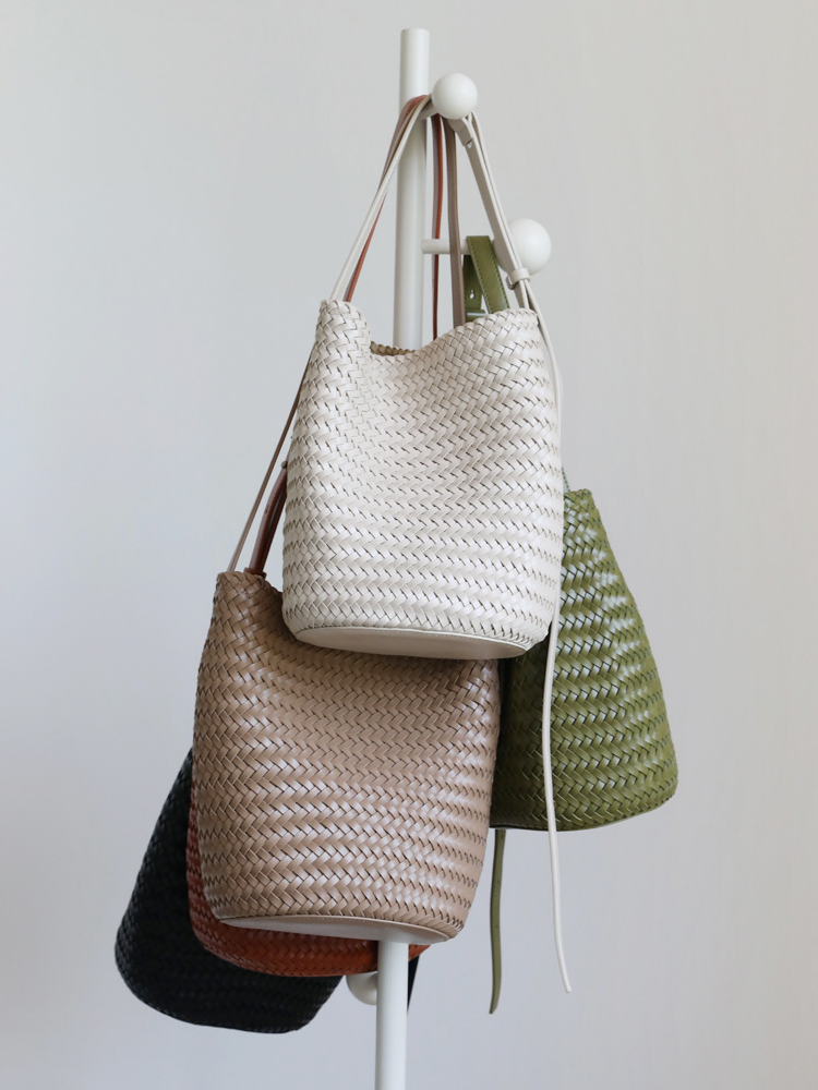 A stylish display of handwoven vegan leather bucket tote bags hanging on a rack, featuring cream, khaki, olive green, black, and brown colorways.
